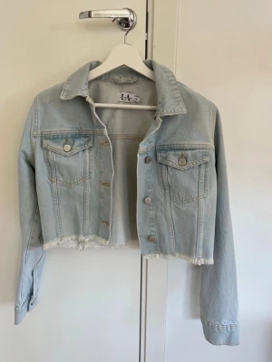 Ljusblå cropped jeansjacka LA - Trendig ljusblå cropped jeansjacka från Linn Ahlborg x NA-KD. Jackan har rå kant nertill, klassisk krage, två bröstfickor med lock och silverfärgade knappar. Perfekt för dig som vill ha en cool och avslappnad look.