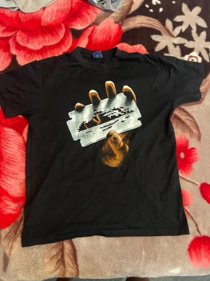 Svart t-shirt corteiz  - Cool svart t-shirt med ett stort tryck av en hand som håller ett rakblad på framsidan. T-shirten har klassisk passform och rund hals. Perfekt för dig som gillar edgy och unika motiv.