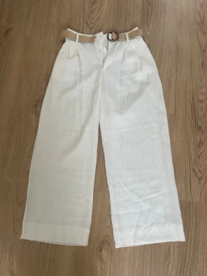 Vita vida linnebyxor med beige bälte - Säljer ett par vita linnebyxor med breda ben och hög midja. Byxorna har ett beige tygbälte med rund metallspänne och praktiska fickor fram. Perfekta för en avslappnad och stilren look. Materialet är luftigt linne som passar bra till sommaren. Aldirg använda💕