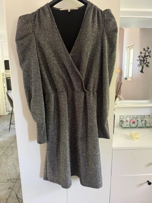 Glittrig omlottklänning med puffärm - Snygg kort glittrig klänning i silver med omlottmodell och markerade puffärmar. Klänningen har v-ringning och långa ärmar, perfekt för dig som vill sticka ut. Materialet är mjukt och följsamt, troligen polyester eller syntet.