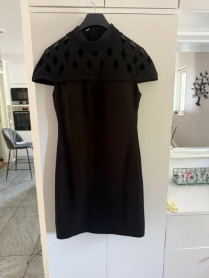 Svart kortärmad klänning med detaljer - Snygg svart klänning med hög hals och korta ärmar. Överdelen har ett unikt rutmönster med utskärningar som ger en cool look. Klänningen är figurnära och slutar ovanför knäna. Perfekt för dig som gillar stilrena plagg med det lilla extra.