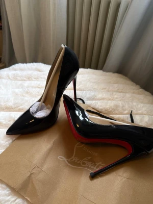 Svarta lackpumps - Säljer ett par klassiska svarta pumps i lack. Skorna har spetsig tå, hög smal klack och den ikoniska röda sulan. Insidan är beige och materialet är glansigt läder. Perfekta för dig som vill ha en elegant och tidlös look. Helt oanvända!