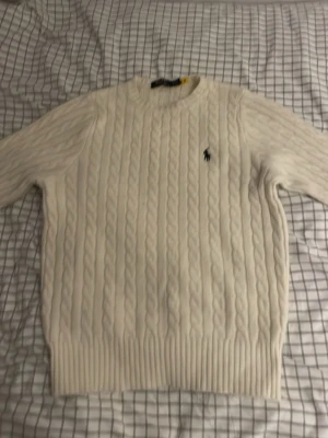 Vit kabelstickad tröja Polo Ralph Lauren - Kabelstickad vit tröja från Polo Ralph Lauren med rund halsringning och den klassiska broderade loggan på bröstet. Tröjan har långa ärmar och ribbade muddar vid ärmslut och nederkant. Perfekt för dig som gillar stilren och tidlös design.