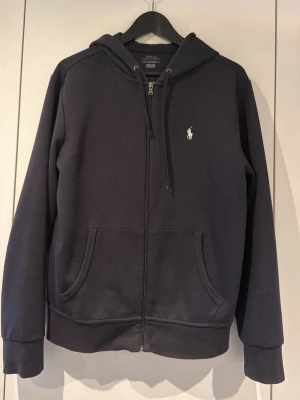 Mörkblå hoodie från Polo Ralph Lauren - Snygg mörkblå hoodie från Polo Ralph Lauren med dragkedja, huva med dragsko och klassisk vit logga på bröstet. Hoodien har känguruficka framtill och ribbade muddar vid ärmar och nederkant. Perfekt för en avslappnad och stilren look.