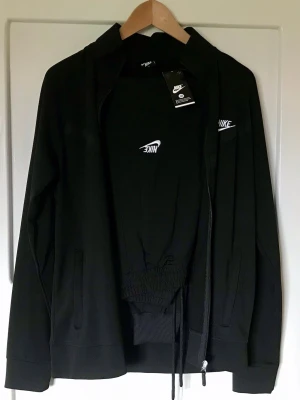 Svart Nike sport set - Nike  set i svart med vit broderad logga på både tröja och byxor. Setet består av en zip-hoodie och matchande byxor med dragsko i midjan. Tillverkat i mjukt och bekvämt material, perfekt för chill dagar hemma.