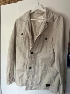 Beige overshirt från H&M - Beige overshirt från H&M i bomull med fyra stora fickor framtill och svarta knappar. Jackan har klassisk krage och lång ärm, perfekt för lager-på-lager och en clean look.