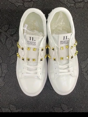 Valentino Rockstud vita sneakers - Snygga vita sneakers från Valentino Garavani med breda band över vristen dekorerade med guldiga nitar. Modellen heter Rockstud Untitled 11 och har en clean design i läder med coola detaljer som sticker ut. Perfekta för dig som vill ha något extra till din outfit.