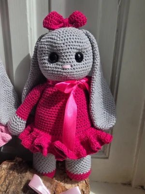 Virkad kanin med rosa klänning - Söt handvirkad kanin i grått med stora svarta ögon, långöron och rosa detaljer. Kaninen har en ceriserosa klänning med volangkant, matchande rosett på huvudet och ett rosa sidenband runt halsen. Perfekt som dekoration eller present. HÖJD CA 28 CM , Virkad i 100 % bomullsgarn och fylld med kuddfyllning i polyester , säkerhetsögon 