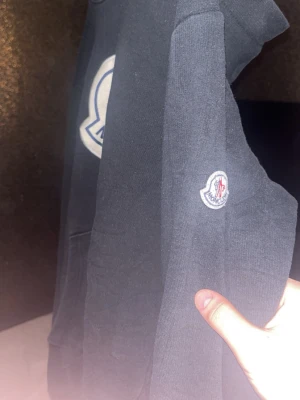 Svart Moncler hoodie med stor logga - Säljer en svart hoodie från Moncler med stor broderad logga på bröstet och en mindre patch på ärmen. Klassisk huva och känguruficka framtill. Perfekt för dig som gillar streetwear och vill ha något stilrent men ändå iögonfallande. Har använts mycket men fortfarande fet.