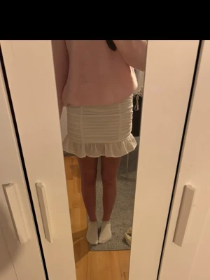 Vit kortkjol med volangkant - Supersöt vit kortkjol med volangkant nertill och rynkade detaljer längs sidorna. Kjolen har en figurnära passform och är perfekt för dig som gillar en feminin och trendig stil. Aldrig använd 