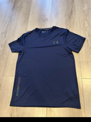 Blå tränings-tshirt från Under Armour - Snygg blå tränings-tshirt från Under Armour med rund hals och korta ärmar. Tillverkad i ett lätt och snabbtorkande funktionsmaterial. Logga i grönt på bröstet och färgglad text längs sidan. Perfekt för gymmet eller löprundan. Känns som M