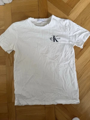 Vit Calvin Klein Jeans t-shirt - Vit t-shirt från Calvin Klein Jeans med klassisk rund hals och diskret CK-logga tryckt på bröstet. Tillverkad i mjuk bomull och har en enkel, stilren design som passar till det mesta. Perfekt basplagg för en clean look.