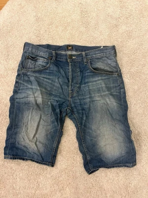 Blå jeansshorts från Lee - Säljer ett par klassiska blå jeansshorts från Lee med fem fickor och knappgylf. Shortsen har en avslappnad passform och slitna detaljer för en cool look. Perfekta till sommaren och lätta att matcha med t-shirt eller hoodie.