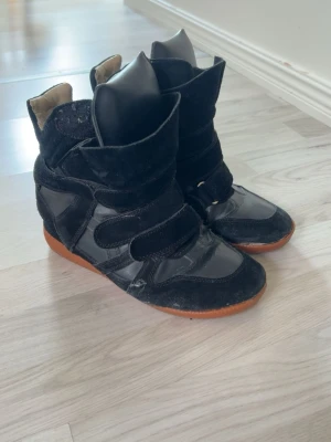 Svarta Isabel Marant wedge sneakers - Snygga svarta wedge sneakers från Isabel Marant med högt skaft och kardborreband. Skorna har en mix av mocka och skinn samt brun sula. Perfekta för dig som vill ha en cool och trendig look med extra höjd. Dom är rätt slitna men fortfarande jättesnygga!💕