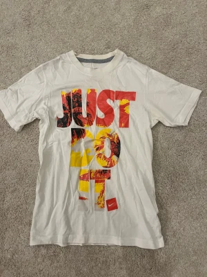 Vit Nike t-shirt med tryck (barn/ungdom)  - Nike t-shirt i vitt med stort färgglatt 'JUST DO IT.' tryck i rött, gult och svart på framsidan. Klassisk rund hals och korta ärmar. Tillverkad i mjuk bomull för en bekväm känsla. Perfekt för dig som gillar sportig och avslappnad stil.                   