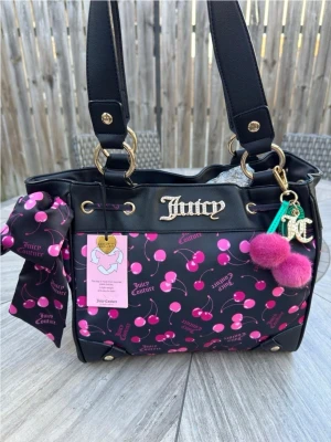 Juicy Couture handväska med körsbär- limited edition  - Juicy Couture handväska i svart konstläder med färgglatt körsbärsmönster och rosa detaljer. Väskan har guldfärgade metalldetaljer, JC-logga framtill, en stor rosett på sidan och fluffiga rosa bollar som nyckelring. Perfekt för dig som gillar statement-accessoarer.