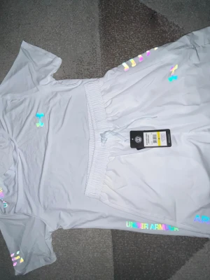 Under Armour Set - Säljer ett vitt träningsset från Under Armour med t-shirt och shorts. Setet har färgglada, reflekterande loggor och detaljer. Materialet är lätt och andas, perfekt för träning eller chill. Elastisk midja på shortsen och klassisk passform på t-shirten.
