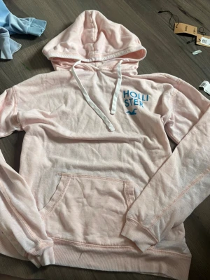 Ljusrosa hoodie från Hollister - Ljusrosa hoodie från Hollister med blå logga och text på bröstet samt stor text 'CALIFORNIA' i blått på ryggen. Klassisk känguruficka framtill, huva med snörning och långa ärmar. Perfekt för en avslappnad och cool stil. Den är tunnare i tyget så det passar jättebra nu på sommaren 