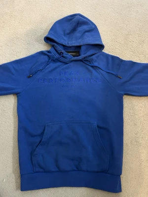 Blå hoodie från Peak Performance - Säljer en blå hoodie från Peak Performance med broderad logga på bröstet. Hoodien har huva med dragsko som är skruvad och går inte och fixa, stor magficka och ribbade muddar vid ärmslut och nederkant. Tillverkad i mjuk bomullsblandning, perfekt för en avslappnad stil.