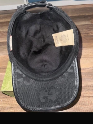 Svart keps från Gucci med GG-mönster - Snygg svart keps från Gucci med det klassiska GG-monogrammet över hela kepsen. Kepsen har böjd skärm och justerbar rem bak. Materialet är en mix av textil och läderdetaljer som ger en exklusiv känsla. Perfekt accessoar för dig som gillar streetwear och lyx. Gucci jumbo