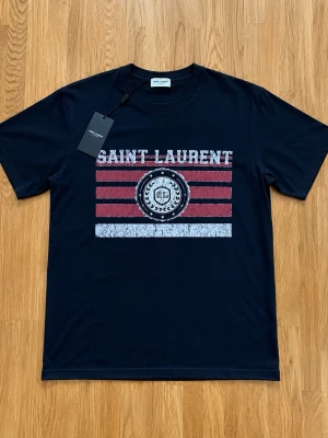 Ysl t-shirt  - Snygg svart t-shirt från Saint Laurent med coolt tryck i vitt och rött framtill. T-shirten har korta ärmar, rund halsringning och är gjord i mjuk bomull. Perfekt för dig som vill ha en trendig och stilren look med exklusiv känsla.