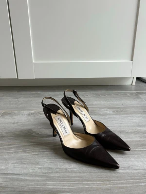 Bruna slingback pumps från Jimmy Choo - Snygga mörkbruna pumps från Jimmy Choo i  läder med spetsig tå och smal klack på runt 10 cm. De är använda fåtal gånger men är tyvärr för små för mig, så i väldigt bra skick. Jättebekväma skor att gå i med justerbar rem bakom hälen. Storlek 35.5 men passar även 36. 