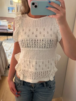 Vit stickad topp med volangdetaljer - Supersöt vit stickad topp med korta ärmar och volangdetaljer längs sidorna. Toppen har ett genombrutet mönster och är croppad i modellen. Perfekt till sommaren.