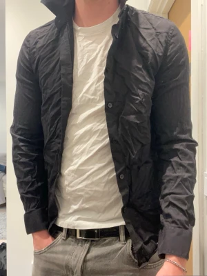 Svart skjorta - Klassisk svart skjorta från H&M No 6 med lång ärm och knappar framtill. Skjortan har en enkel, stilren design och är tillverkad i bomull vilket gör den bekväm och lätt att bära. Perfekt för dig som gillar en clean och tidlös look.