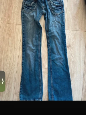 Blå bootcut jeans med detaljer - Säljer ett par blå bootcut jeans med snygga slitningar och unika fickdetaljer, inklusive dragkedjor på bakfickorna och en liten röd patch. Klassisk femficksmodell i mjuk denim med breda hällor och cool tvätt.