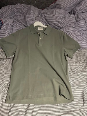 Grön pikétröja från GANT - Snygg grön pikétröja från GANT i regular fit. Klassisk modell med krage, två knappar och diskret GANT-logga broderad på bröstet. Tillverkad i ett mjukt och bekvämt bomullsmaterial som passar perfekt till jeans eller shorts. Helt oanvänd 