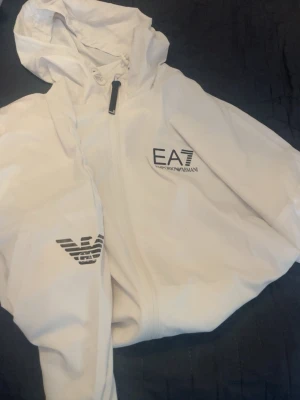 EA7 tracksuit - Snygg vit vindjacka från Emporio Armani EA7 med huva och dragkedja framtill. Jackan har logga på bröstet och ärmen, samt minimalistisk design. Perfekt för dig som gillar stilrena och sportiga plagg.