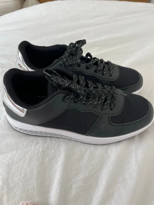 Oanvända sneakers - Nya svarta sneakers med silvriga detaljer på hälen och vita sulor. Storlek 41, men små i storleken. Passar för den som har 39