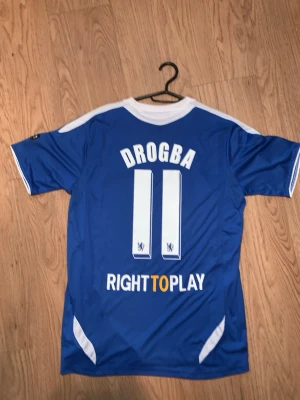Chelsea Drogba matchtröja blå - Chelsea FC matchtröja i blått med vita detaljer, Adidas-logga och Samsung som sponsor. Drogba och nummer 11 på ryggen, samt Champions League-märke på ärmen. Tillverkad i lätt och ventilerande material, perfekt för fotboll eller samlare.