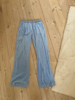 Juicy couture - Säljer ett par ljusblå mjukisbyxor i mjuk velour med raka ben och bred resår i midjan. Byxorna har två stora fickor framtill med dekorativa guldknappar. Perfekta för en chill och avslappnad stil.
