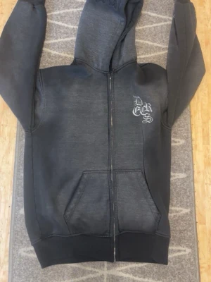 Mörkgrå zip hoodie med tryck - Säljer en mörkgrå hoodie med dragkedja från DCS. Hoodien har huva, fickor framtill och ett vitt tryck på bröstet. Perfekt för en avslappnad streetwear-look. Materialet känns mjukt och bekvämt, och passformen är klassisk med ribbade muddar.