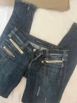 MissSixty jeans med dragkedjor - Snygga mörkblå jeans från Miss Sixty med slitningar och coola silverfärgade dragkedjor framtill. Jeansen har låg midja, fem fickor och detaljerade sömmar. Perfekta för en trendig och edgy look.