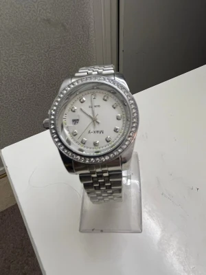 Silverfärgad klocka från MAX-Y - Stilren klocka från MAX-Y i silverfärg med metallarmband och vit urtavla. Klockan har glittrande stenar runt boetten och markerade timindex med stenar. Klassisk design som passar till många stilar.