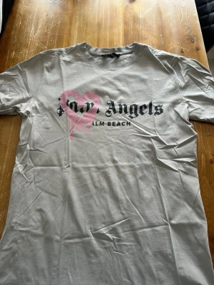 Palm Angels t-shirt med rosa hjärta - Snygg grå t-shirt från Palm Angels med svart text och ett stort rosa hjärta med drip-effekt på bröstet. Klassisk rund hals och korta ärmar. Perfekt för dig som gillar streetwear och vill sticka ut med en unik design.