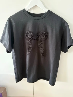 Svart t-shirt med rosettdetalj - Snygg svart t-shirt med en stor dekorativ rosett framtill i matchande tyg. T-shirten har klassisk passform och rund halsringning. Perfekt för dig som vill ha en enkel men ändå unik look.