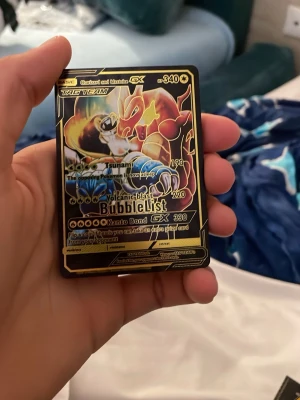 Pokémonkort Charizard & Blastoise GX - Säljer ett Pokémonkort med Charizard och Blastoise GX i tag team-version. Kortet har en svart och guldig design med holografiska detaljer och tydliga attacker som Tsunami och Volcanic Blast. Perfekt för samlare eller Pokémonfans.