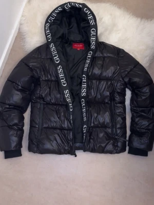 Svart pufferjacka från Guess - Snygg svart pufferjacka från Guess med stor huva och logotyptext längs kanten. Jackan har en glansig finish, quiltad design och muddar vid ärmsluten. Perfekt för kalla vinterdagar och riktigt trendig look.