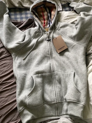 Grå zip hoodie från Burberry - Snygg grå zip hoodie från Burberry med klassiskt rutigt foder i huvan och dragsko. Hoodien har fickor framtill och ribbade muddar vid ärmar och nederkant. Perfekt för en chill och stilren look.