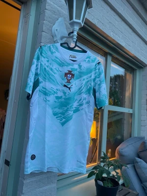 Portugal fotbollströja Puma turkos - Säljer en Portugal fotbollströja från Puma i vitt och turkost med vågigt mönster. Tröjan har korta ärmar, rund hals och Portugal-emblemet på bröstet. Tillverkad i lätt och ventilerande material, perfekt för träning eller match.