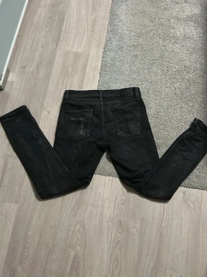 Svarta lean dean jeans från nudie jeans - Säljer ett par svarta lean dean jeans från nudie jeans med klassisk femficksdesign och smal passform. Jeansen har svarta sömmar och är tillverkade i stretchigt denim för extra komfort. Perfekta för en stilren och modern look.