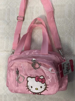 Hello Kitty rosa Axelväska / handväska - Söt rosa handväska med Hello Kitty-motiv från Sanrio. Väskan har både handtag och justerbar axelrem, flera fack med dragkedjor och detaljer i form av Hello Kitty-berlocker. Tillverkad i glansigt syntetmaterial med mönster av Hello Kitty och små rosetter.