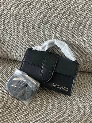 Svart Jacquemus Le Chiquito handväska - Säljer en svart Jacquemus Le Chiquito handväska i slätt läder med silver logotyp framtill. Väskan har ett topphandtag och magnetlås, samt medföljande axelrem. Perfekt för dig som gillar trendiga och stilrena accessoarer. Made in italy,dustbag kommer med