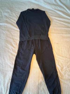 Mörkblå loungewear-set från Hugo Boss - Mysigt mörkblått loungewear-set från Hugo Boss med sweatshirt och matchande byxor. Tröjan har rund halsringning och broderad logga framtill. Byxorna har dragsko i midjan och ribbade muddar vid benslut. Perfekt för chill dagar hemma.