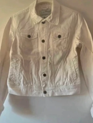 Vit jeansjacka med  knappar som ny - Snygg vit jeansjacka med klassisk krage och bruna metallknappar framtill. Två bröstfickor med lock och knapp samt två sidofickor. Rak passform och långa ärmar. Perfekt att slänga över en t-shirt för en clean look. Storlek 34, men stor i storleken och kan även passa 36. Hade på mig bara 2 gånger.