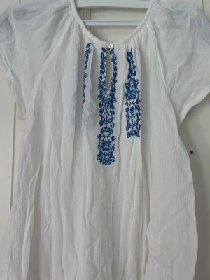 Vit blus med blå brodyr KappAhl - Vit blus från KappAhl med korta ärmar och blå broderade detaljer framtill. Blusen har en lätt och luftig känsla, rund halsringning med liten knapp och är gjord i ett tunt, vävt material. Perfekt för en avslappnad och stilren look.
