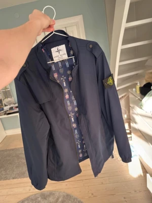 Mörkblå vindjacka från Stone Island - Snygg mörkblå vindjacka från Stone Island med klassisk patch på vänster ärm. Jackan har dragkedja framtill, hög krage och coolt mönstrat innerfoder. Perfekt för dig som vill ha en stilren och trendig look.